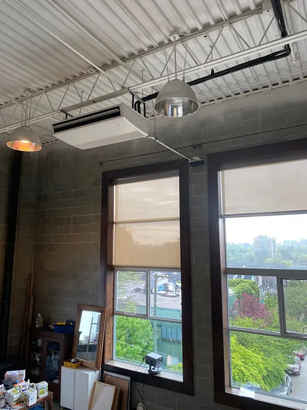The same commercial interior in portrait framing: a ceiling-suspended fan-coil unit and its line set against a block wall, with tall timber-framed windows below.