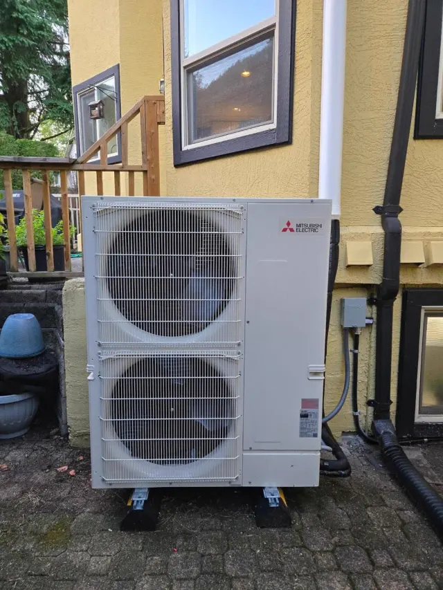 A twin-fan outdoor unit standing on pads beside a house wall, next to a wooden deck, with the refrigerant lines and drain routed into the wall behind it.