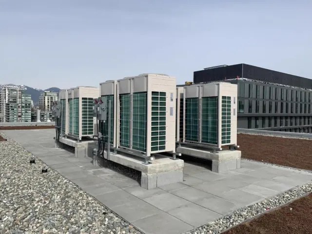 A row of outdoor heat pump modules set on plinths across a flat roof, with the city and the mountains behind.